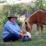 Judith wears a blue shirt and wide-brimmed hat. She sits on the ground next to a light brown miniature horse and a Jack Russell dog.und next to a