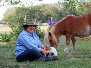 Judith wears a blue shirt and wide-brimmed hat. She sits on the ground next to a light brown miniature horse and a Jack Russell dog.und next to a